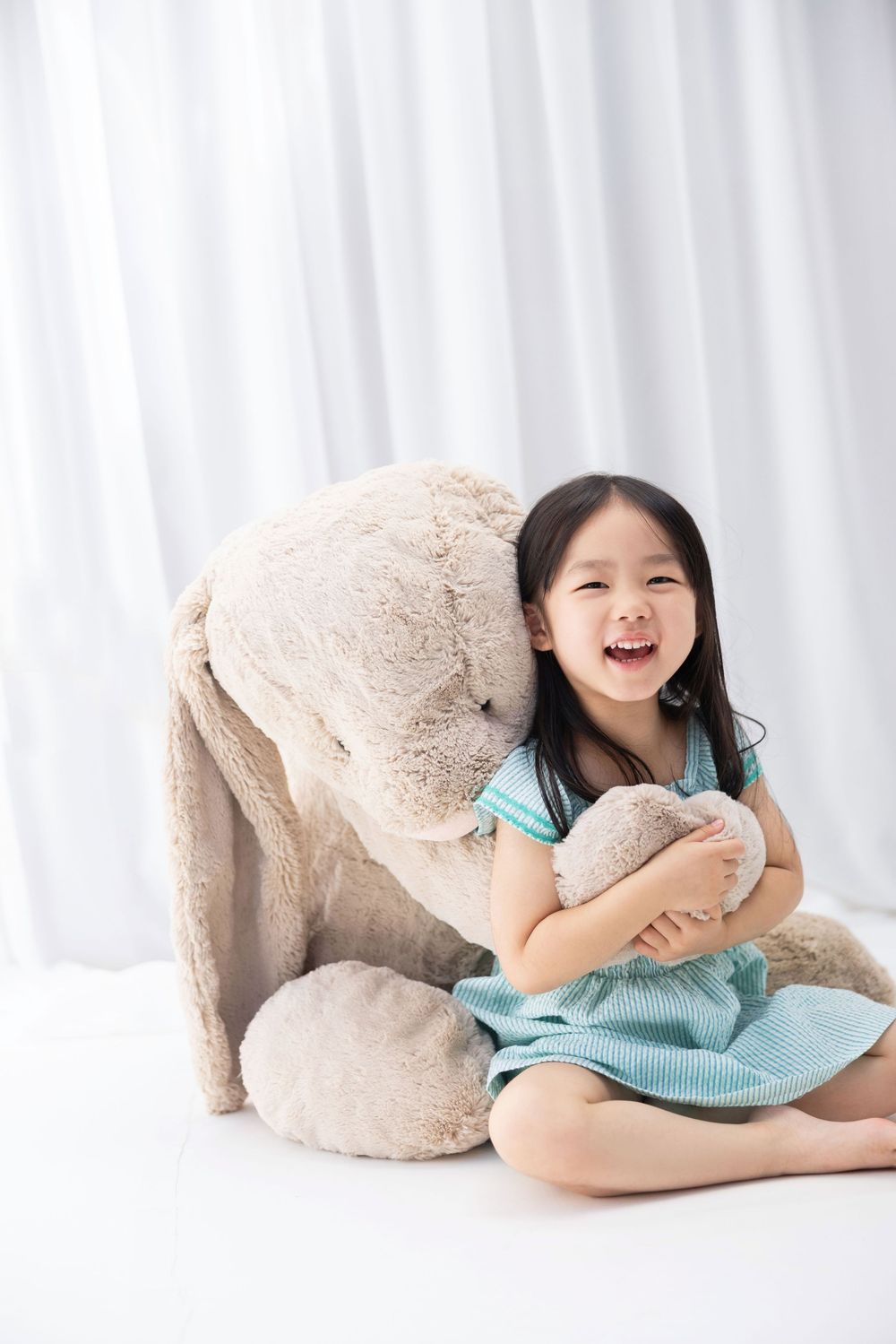 A child in a blue dress sits and laughs while hugging a large plush stuffed bunny against a white curtained background.