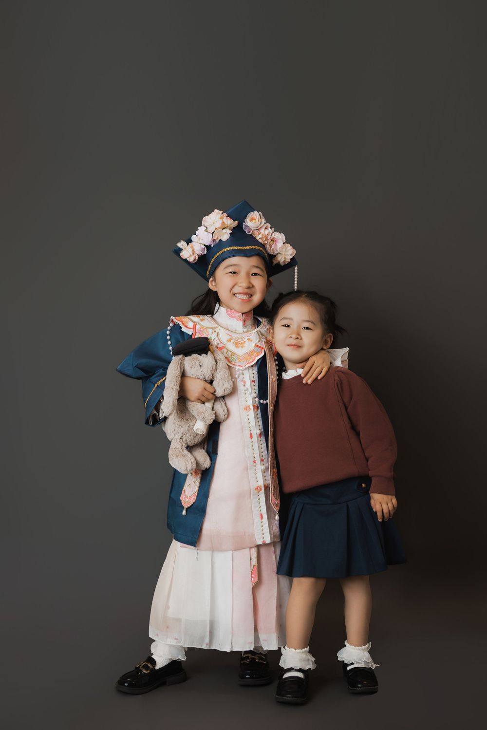 Two young Asian girls posing together, one in traditional Chinese costume with floral headpiece holding a stuffed toy.