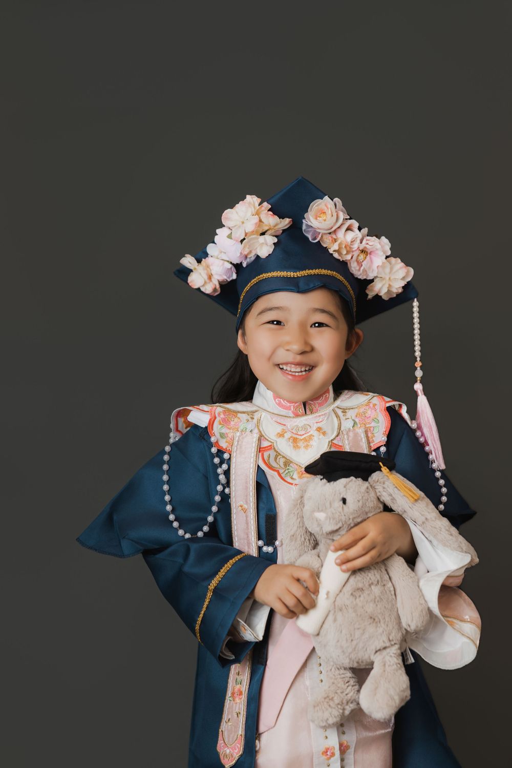 Smiling Asian girl in graduation gown and floral cap holding a stuffed bunny toy, studio portrait.