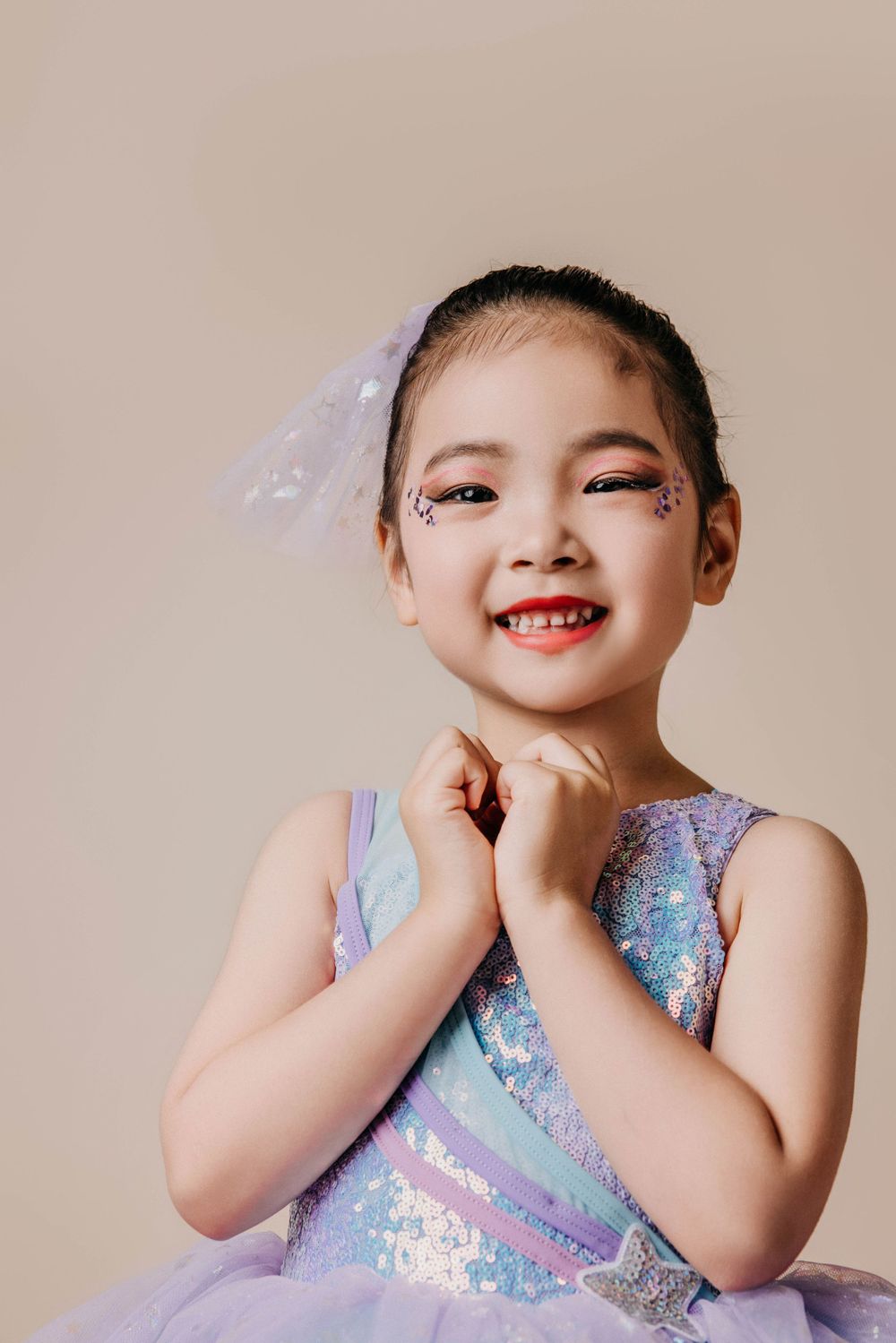 Young dancer wearing sparkly purple tulle dress and face glitter poses with cheerful expression.
