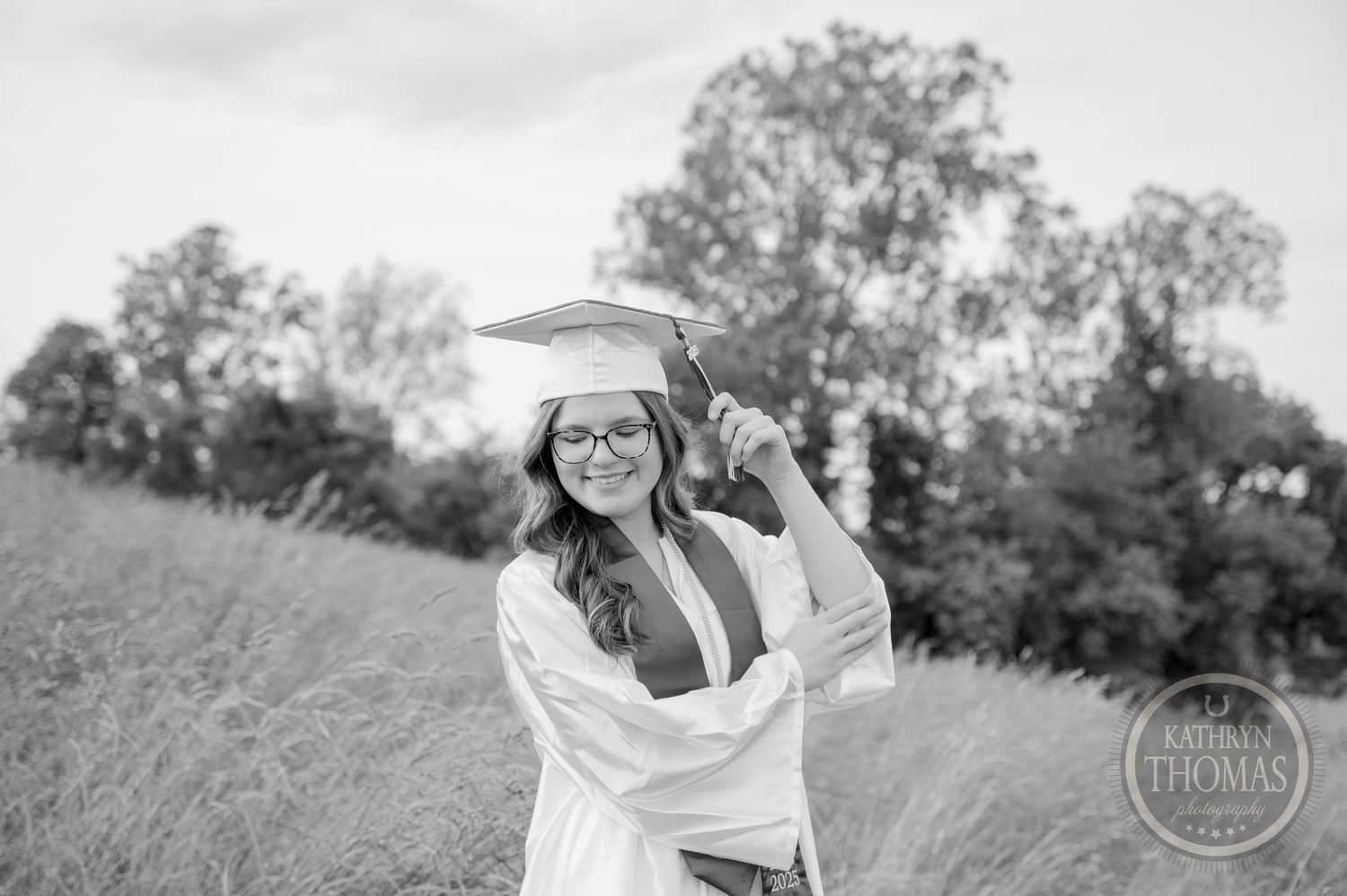 Monochrome photo of graduate celebrating in cap and gown while standing in grassy field with trees in background.