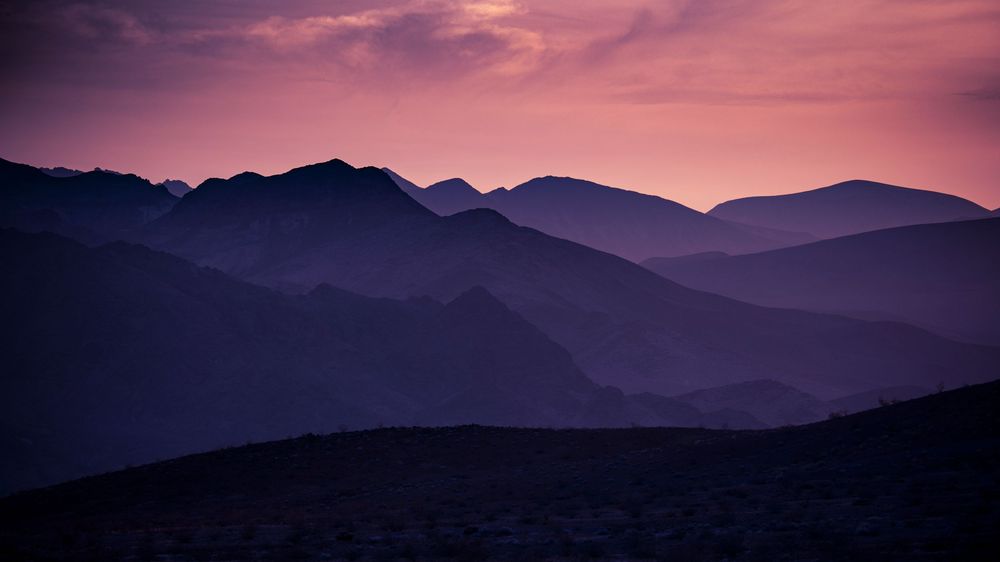 Serene purple and pink sunset casting soft hues over layered silhouetted mountain ridges.