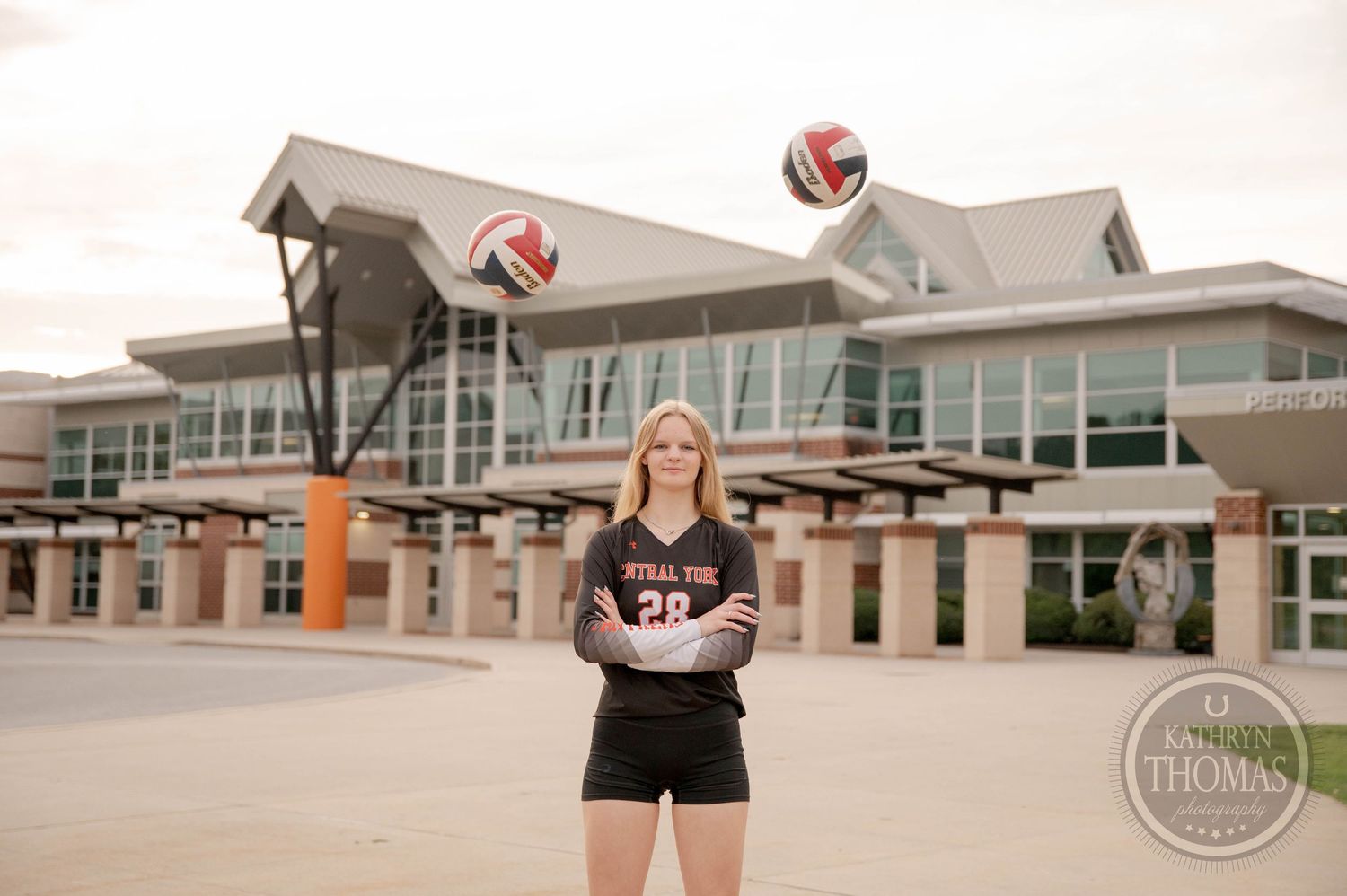 A volleyball athlete stands in front of a modern sports facility with volleyballs floating in the edited photograph.