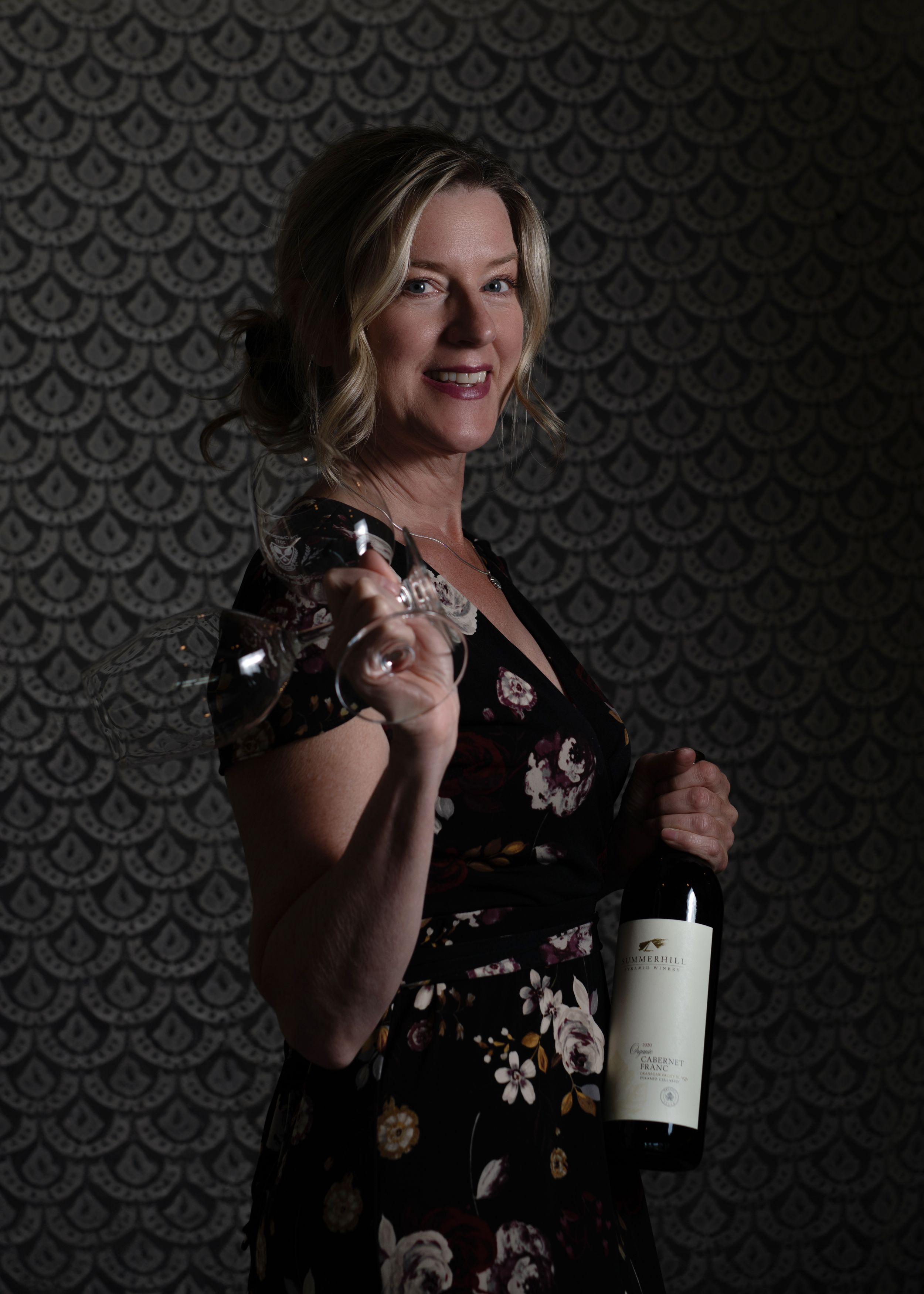 Self-portrait of Tania Veitch in black floral dress holding Summerhill wine bottle and glasses, smiling before lace backdrop.