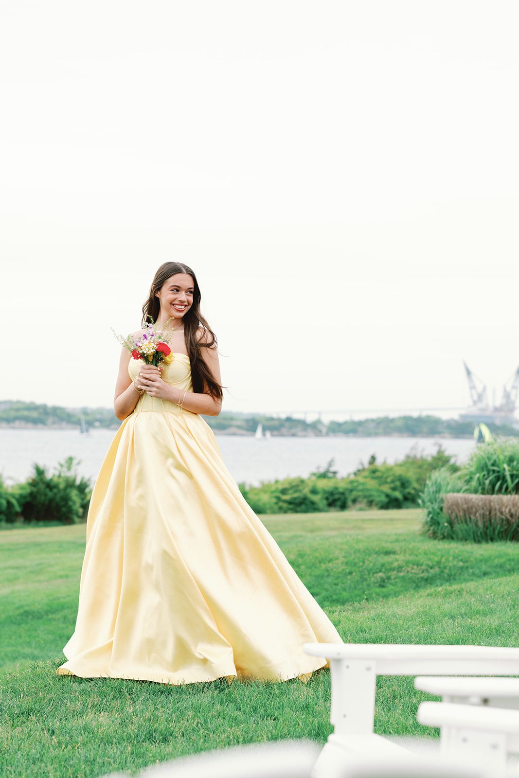 Woman in flowing yellow gown twirls joyfully in outdoor garden setting near waterfront on sunny day.