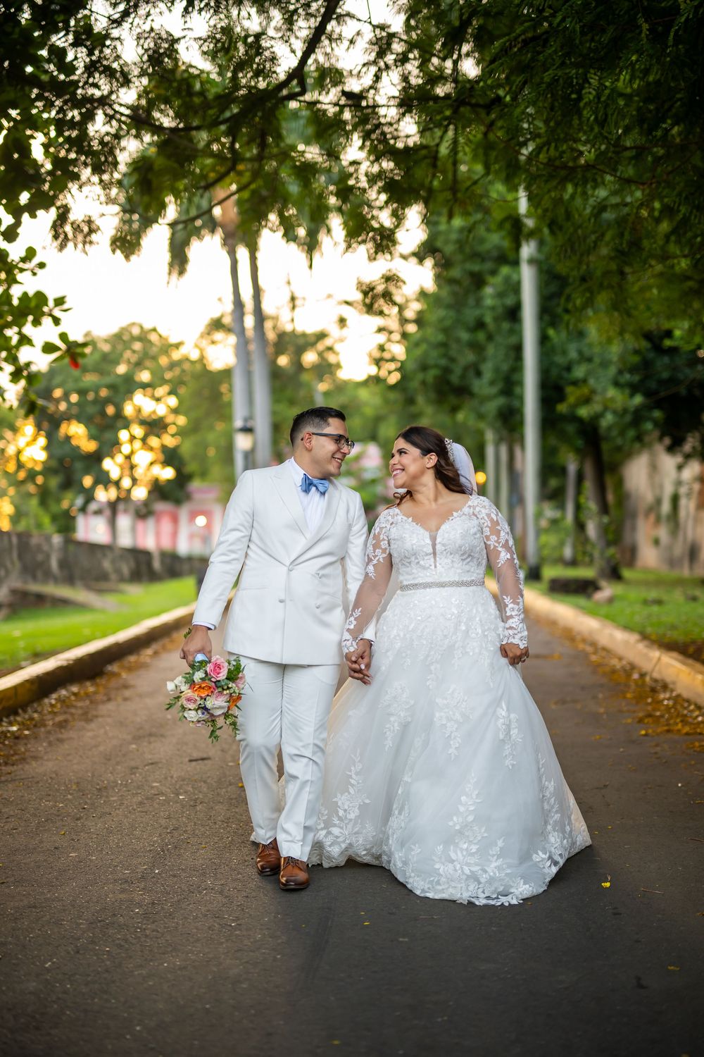 Wedding couple strolls along a tree-lined path at sunset with vibrant bouquet of flowers.