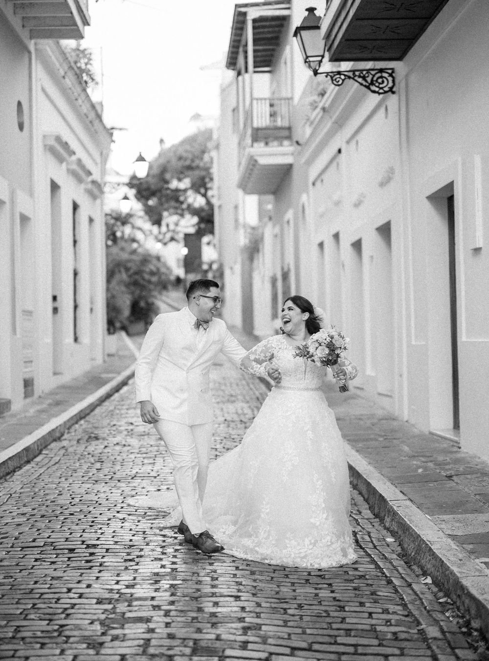 A couple in wedding attire walks down a cobblestone street between white colonial buildings in Old San Juan.