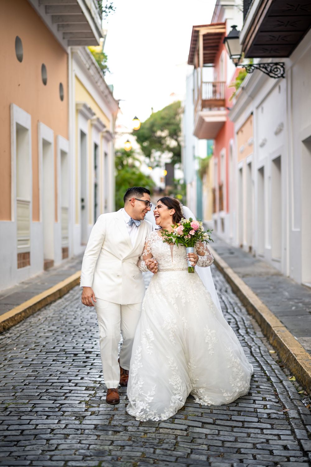 A couple in white wedding attire walks together on a cobblestone street lined with colorful buildings in Old San Juan.