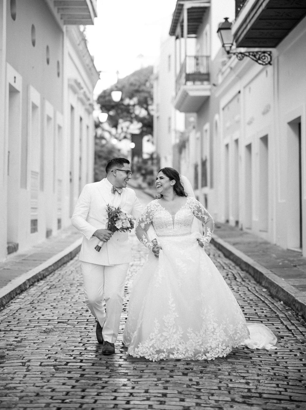 A couple walks down a cobblestone street in Old San Juan, dressed in white wedding attire with historic buildings behind them.
