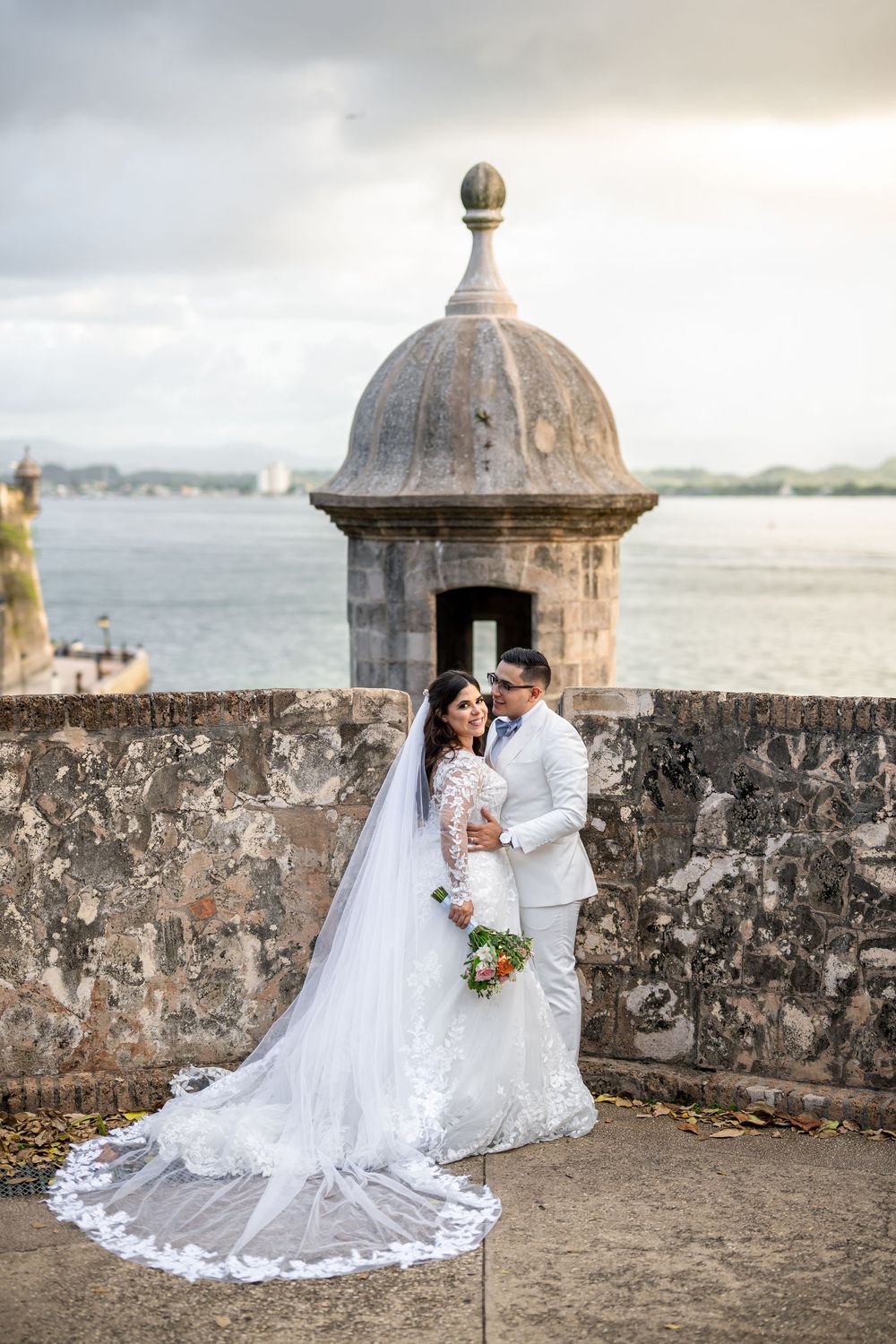 A couple in wedding attire embrace by a historic stone watchtower overlooking a waterfront vista.