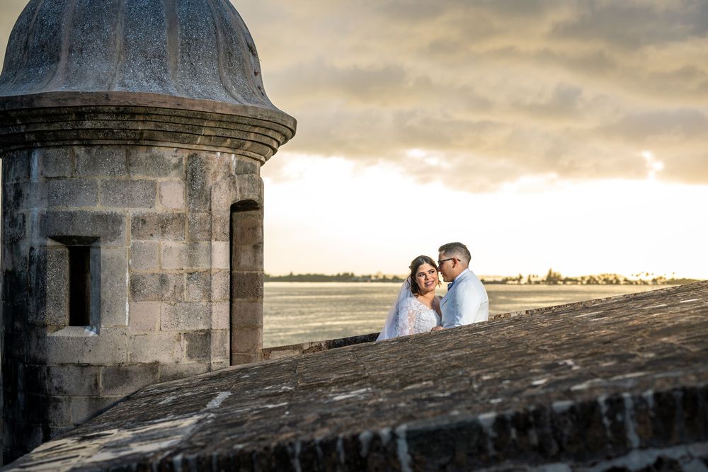 Romantic couple shares an intimate moment at sunset near a historic stone building with dramatic cloudy sky backdrop.