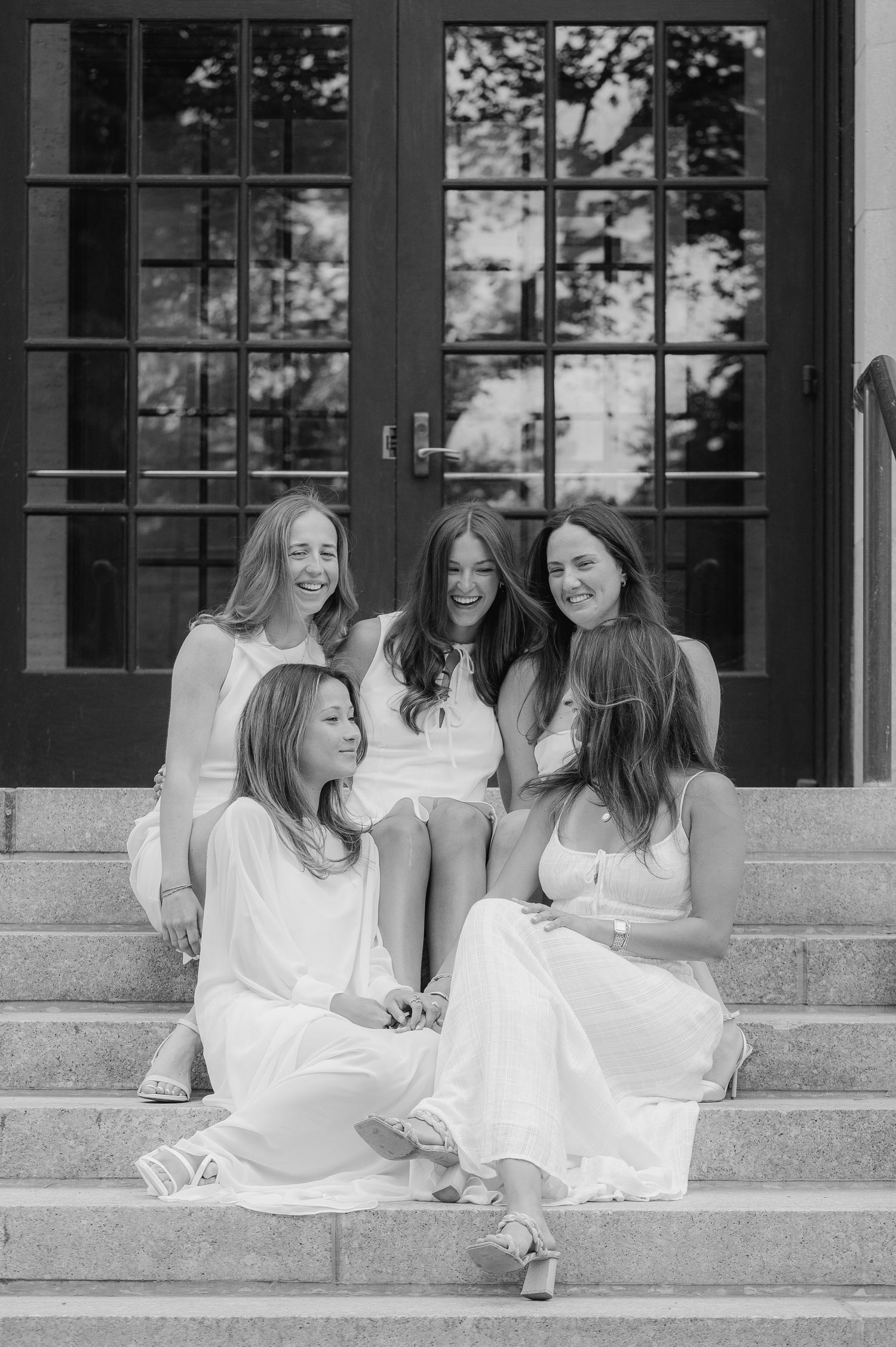 Five young women in white dresses pose together on stone steps in a black and white photo.
