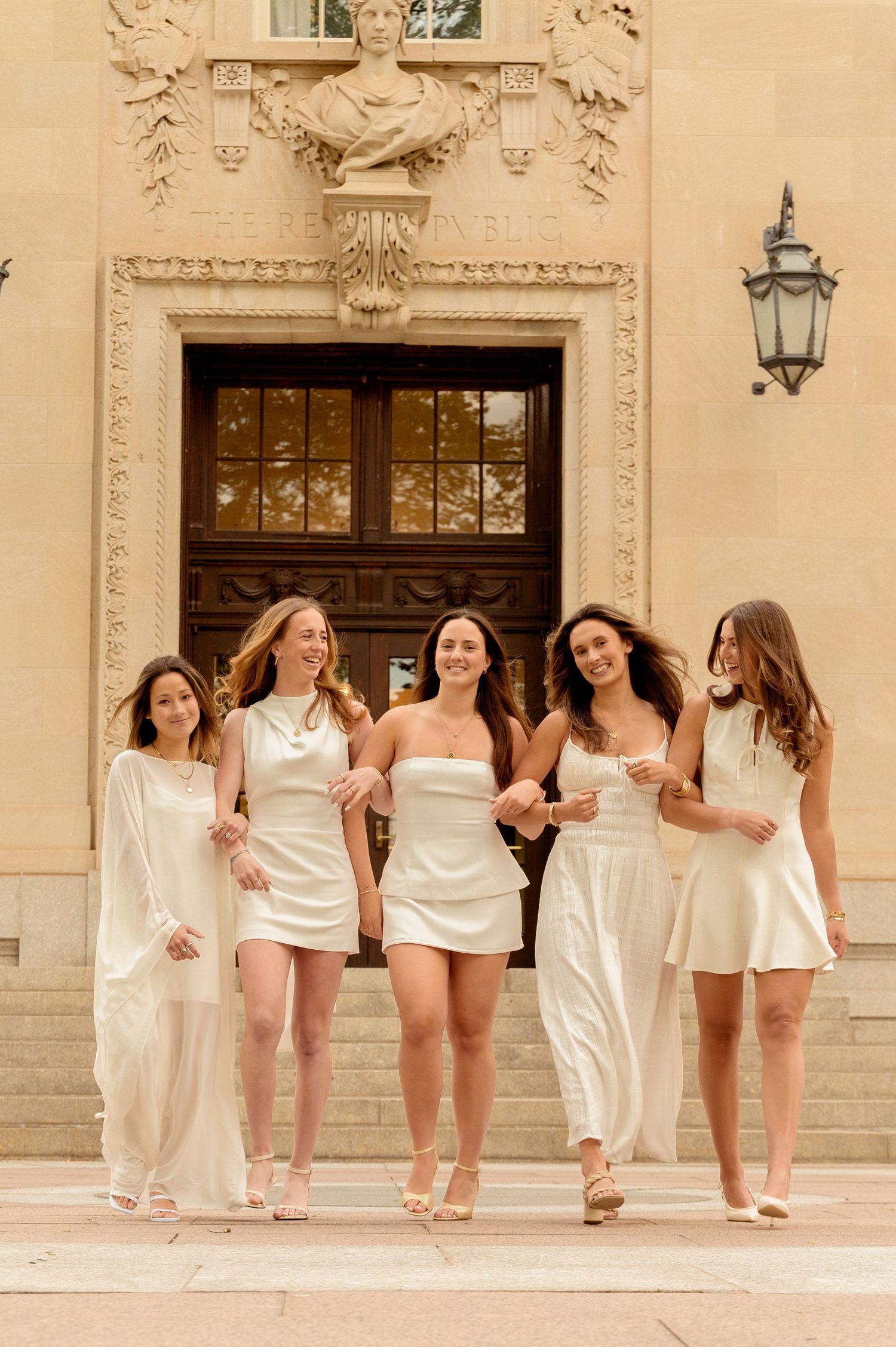 Five women in white dresses walk together smiling in front of an ornate building entrance.
