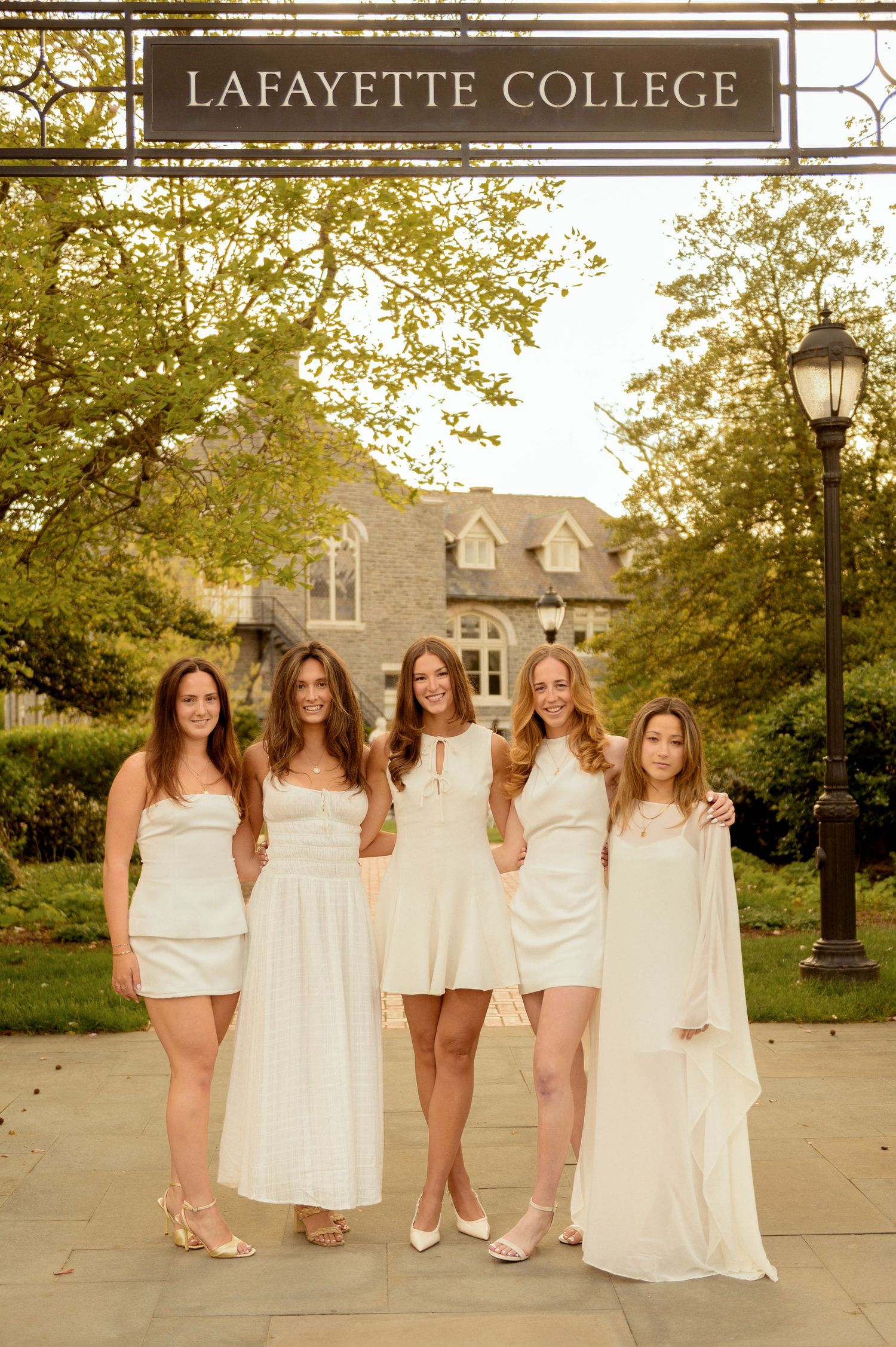 Five women in white dresses posing under Lafayette College sign on campus.