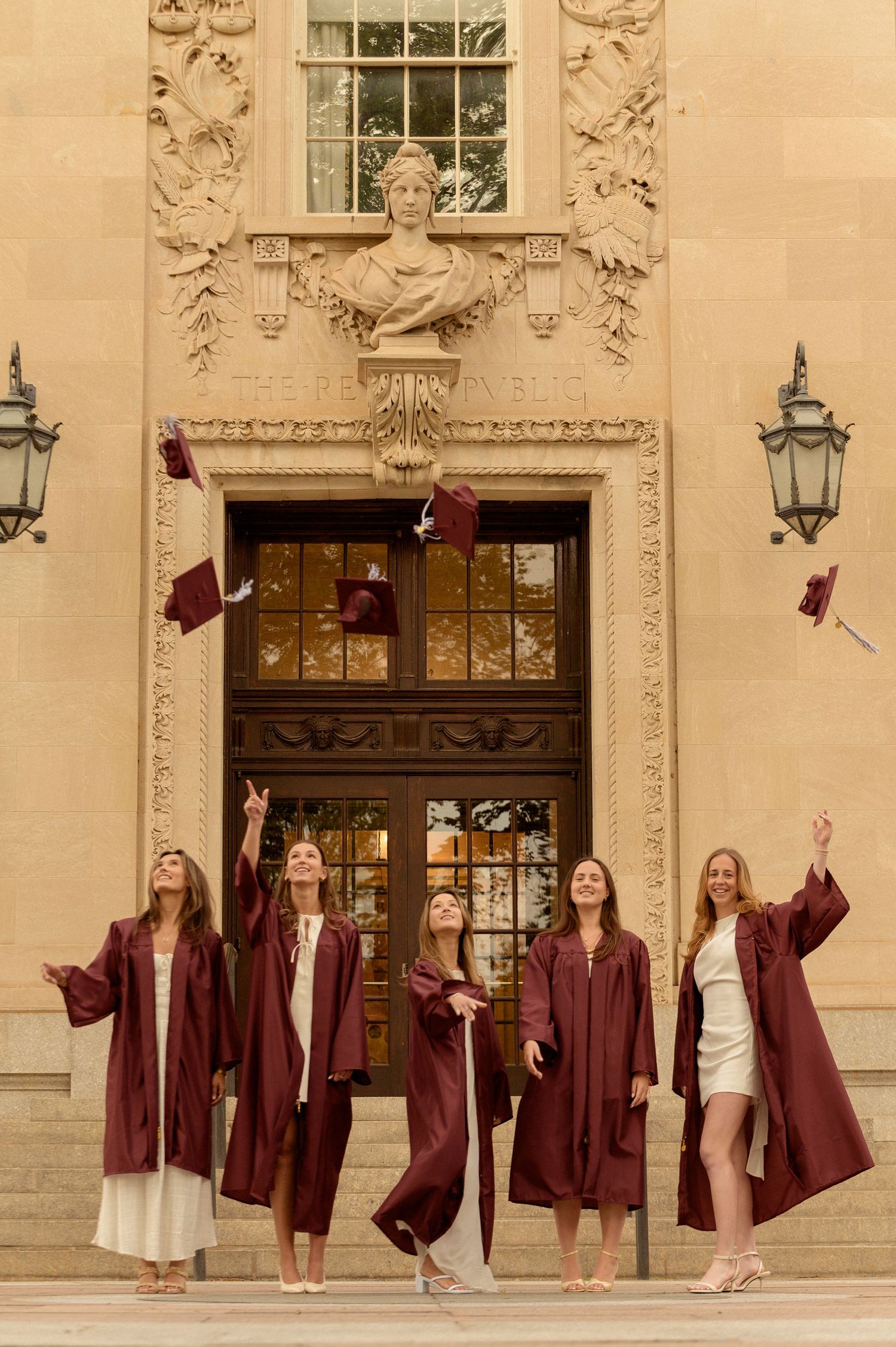 Five graduates in maroon robes tossing caps in front of ornate university building.