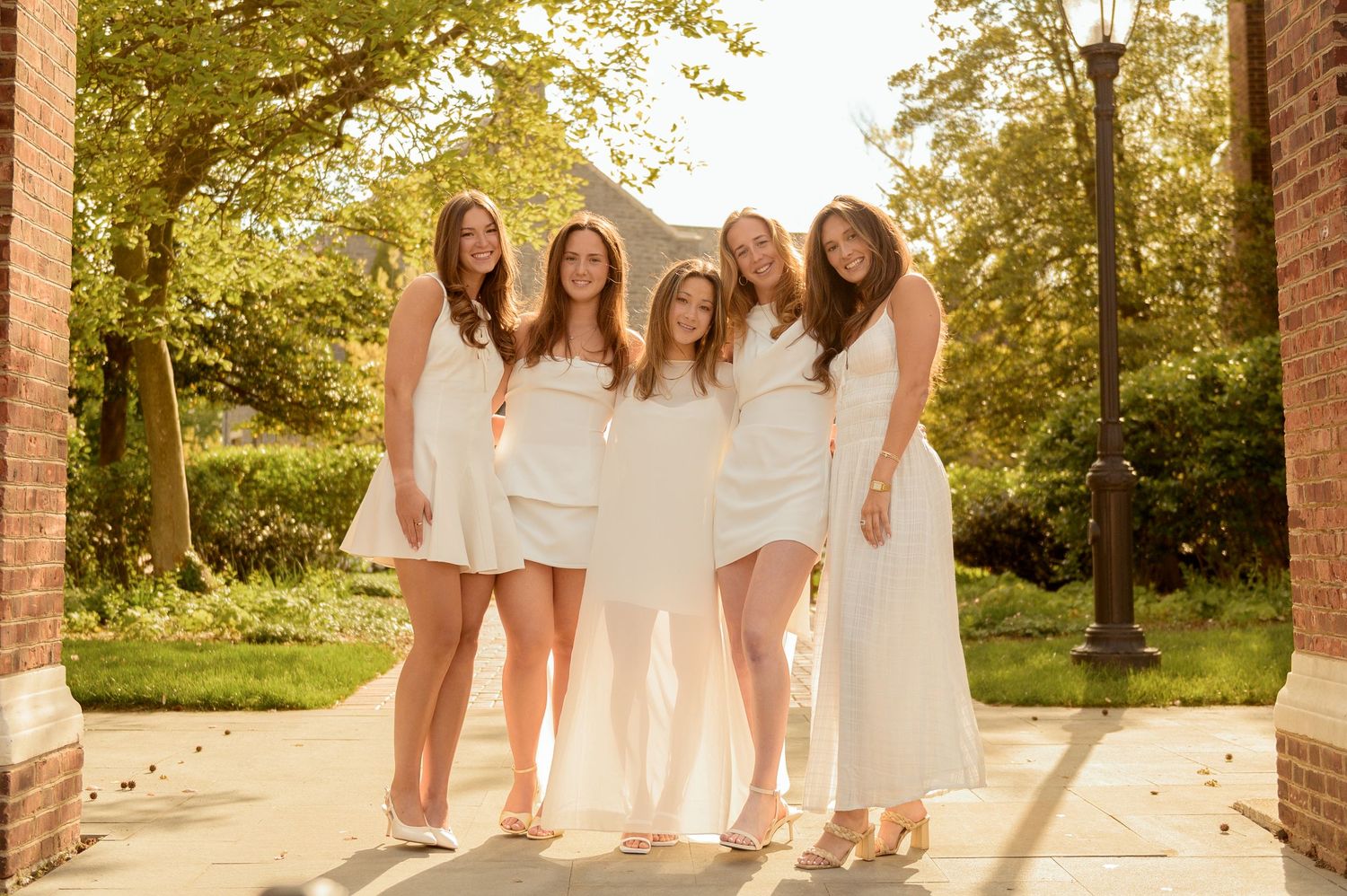Five young women in white dresses smiling together outdoors on a sunny day.