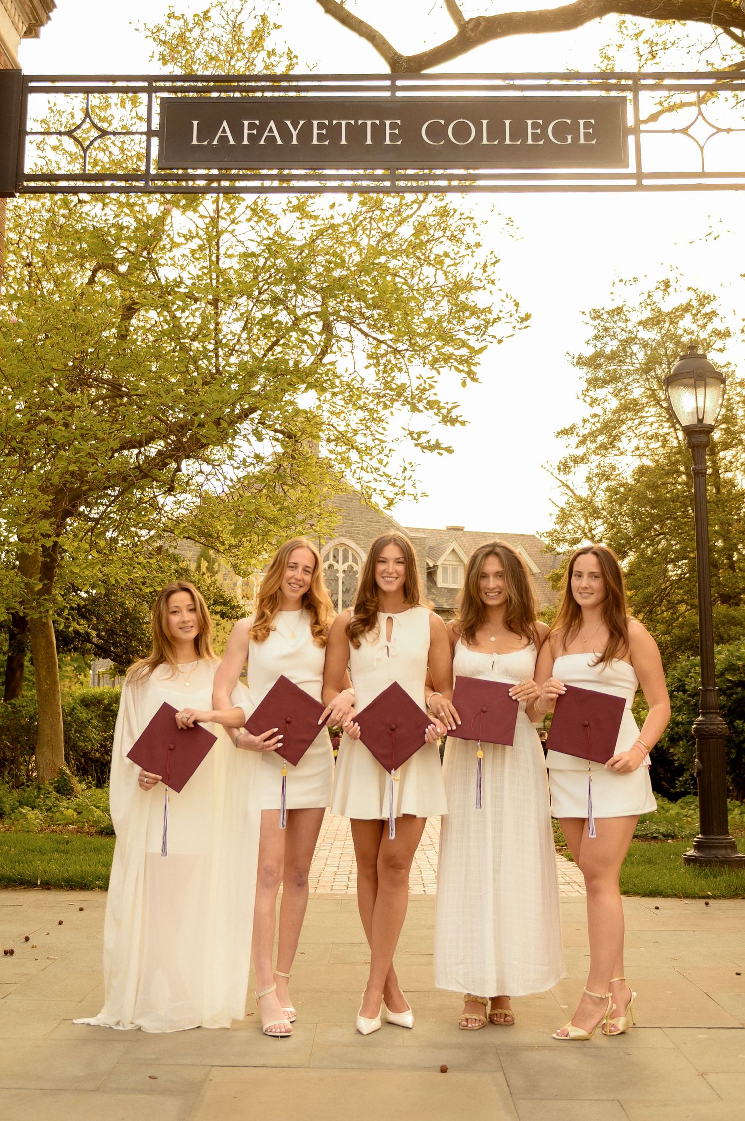 Five Lafayette College graduates in white dresses holding maroon caps and diplomas under the campus sign.