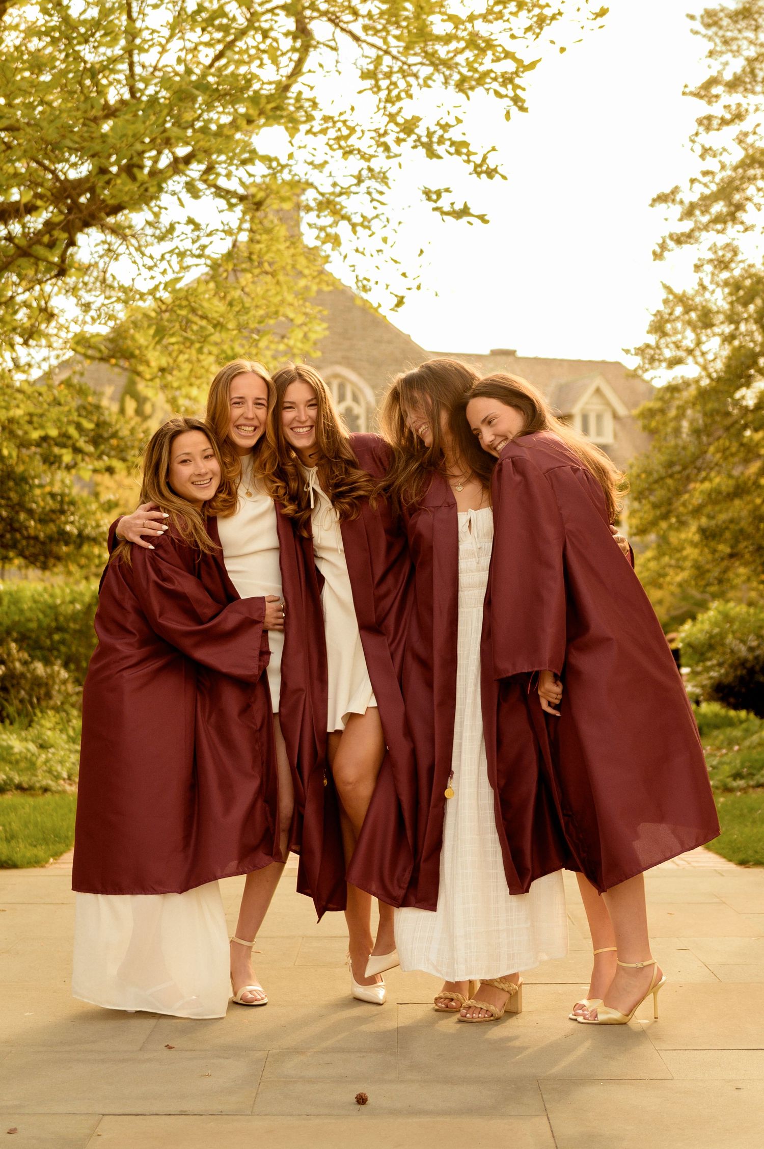 Four young women in maroon graduation gowns and white dresses pose together outdoors on a campus walkway.