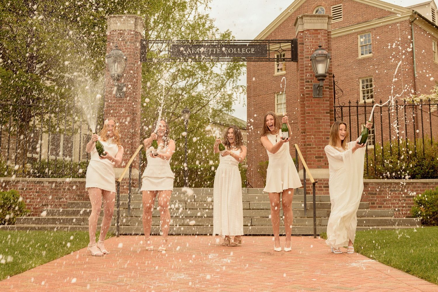 Five women in white dresses celebrate outdoors at Lafayette College, tossing confetti in a joyful graduation photo.