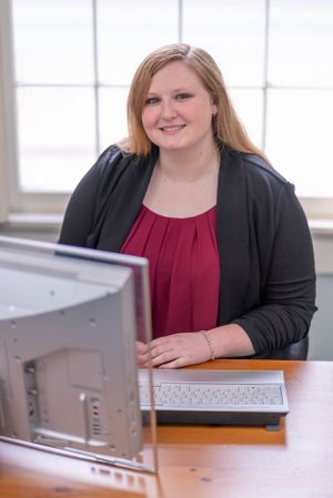 A professional headshot of someone wearing a burgundy top and black blazer at a desk.