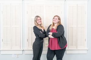 Two people in casual business attire laughing and posing together in front of a white wall.