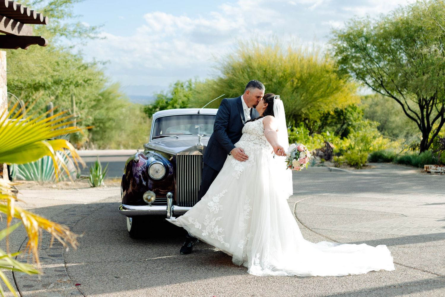 A romantic wedding couple poses next to a vintage Rolls Royce car with desert landscape in the background.
