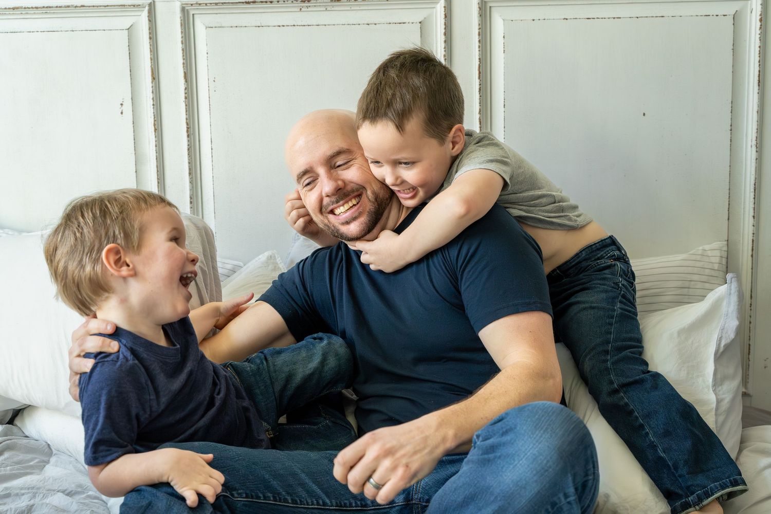 Playful family moment filled with laughter and hugs while relaxing together on a white bed. In-home photography session.