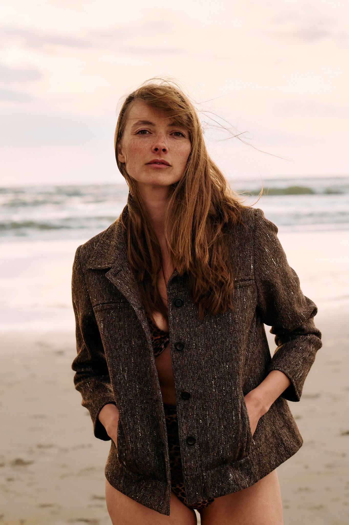 A model wearing a tweed jacket poses on a beach during an overcast day with waves in the background.