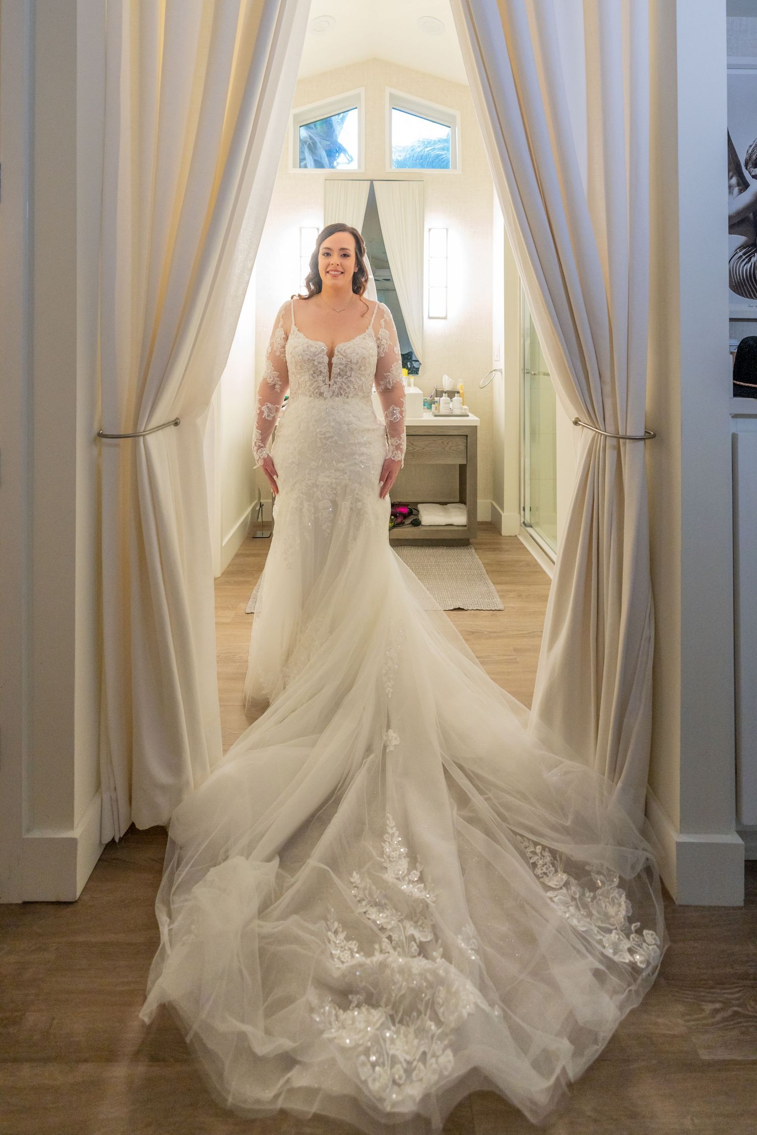 Elegant wedding dress with intricate lace details and flowing train framed by white curtains.
