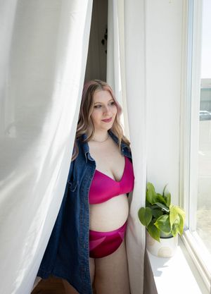 In studio, maternity portrait with pregnant woman wearing pink lingerie looking out the window.