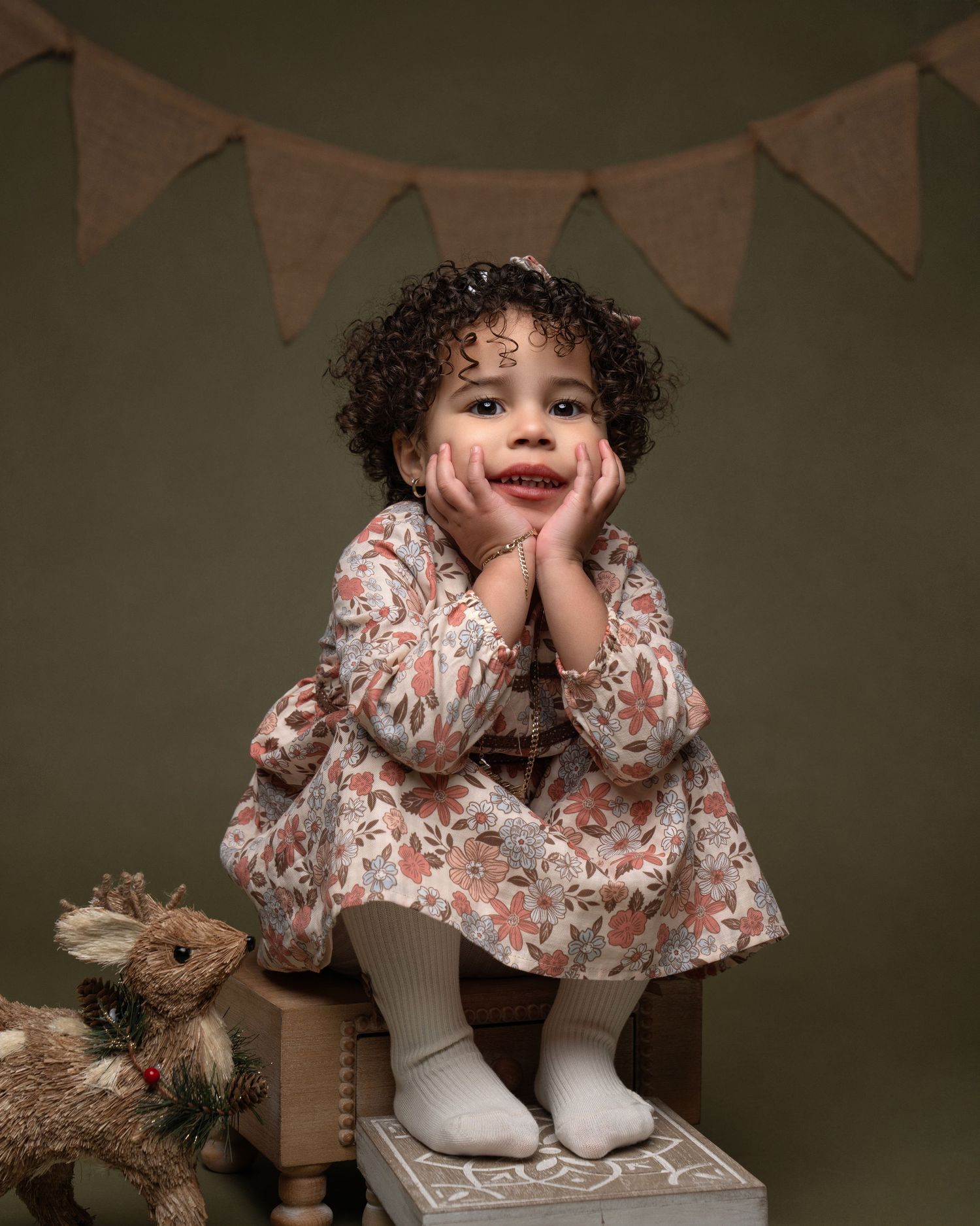 A child in a floral dress sits on a wooden stool with bunting decorations in the background.