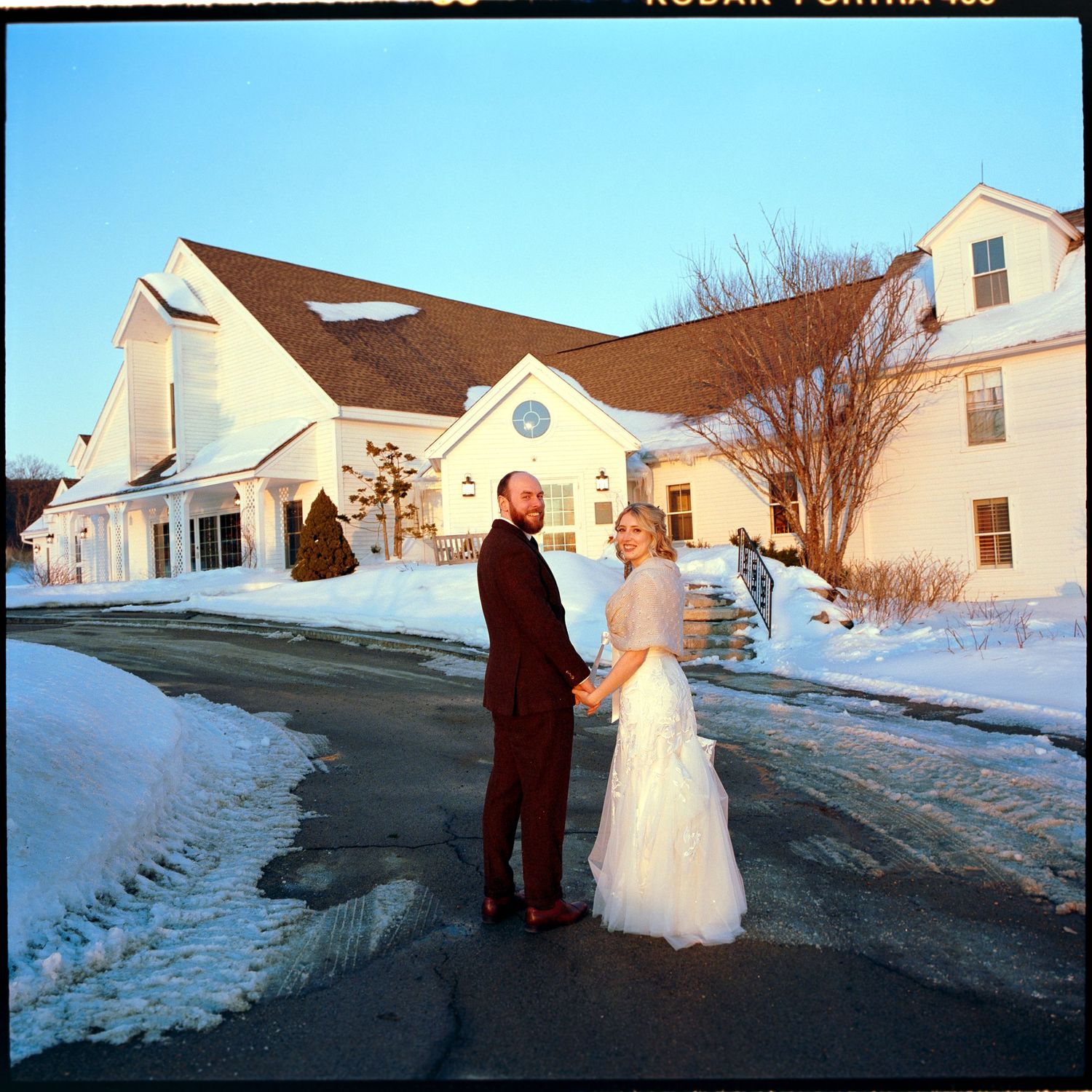 A winter wedding scene with a couple standing in a driveway outside a snowy white church or event venue.