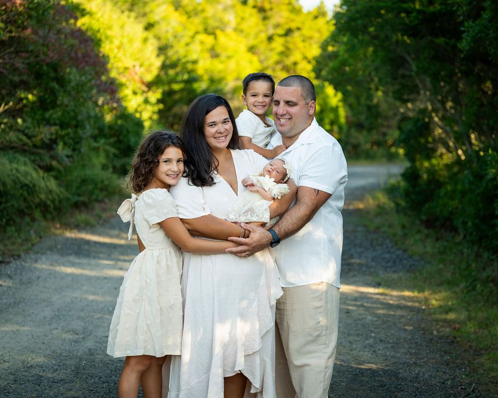 Family of five posing on tree-lined path with newborn baby outdoors.