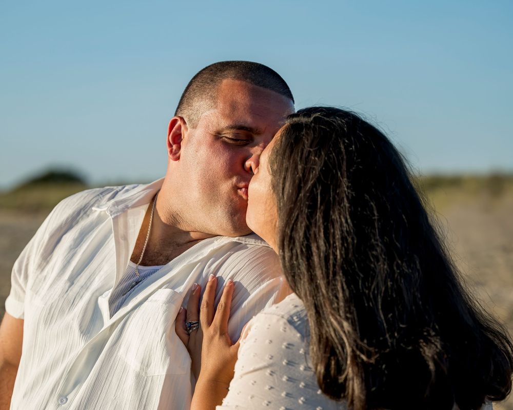 Couple sharing a romantic kiss on sandy beach in white outfits.