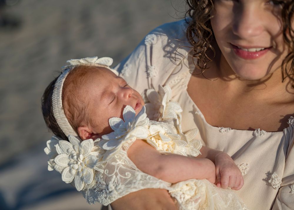 Mother in white dress cradling sleeping newborn baby with floral headband.