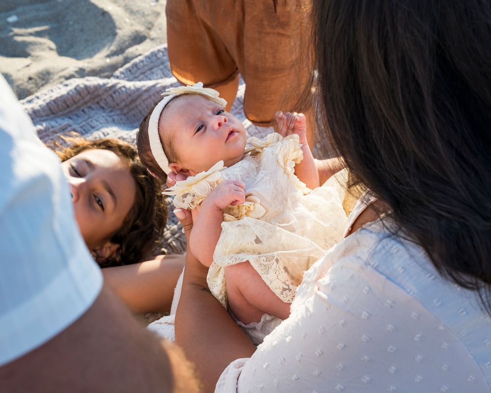 Family gathered around newborn baby lying on blanket at sunny beach.