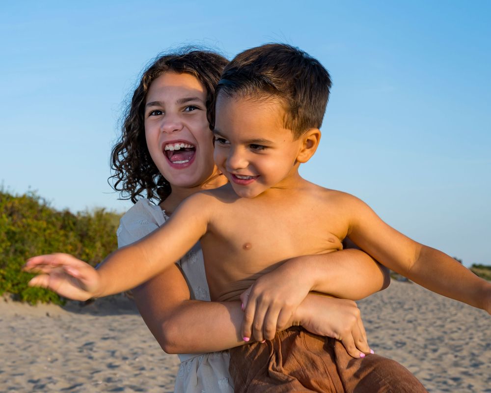 Two happy children playing on a sandy beach, with a girl hugging a shirtless boy from behind under a clear blue sky.