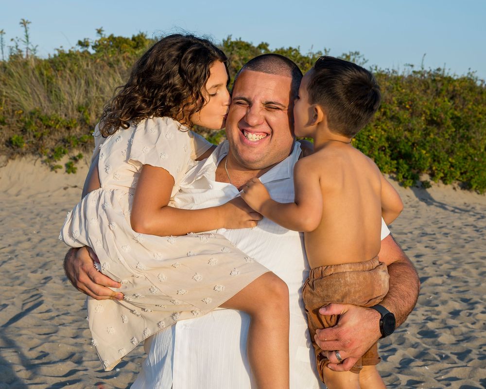 A smiling man holds two young children on a sandy beach as they both lean in to kiss his cheeks at sunset.