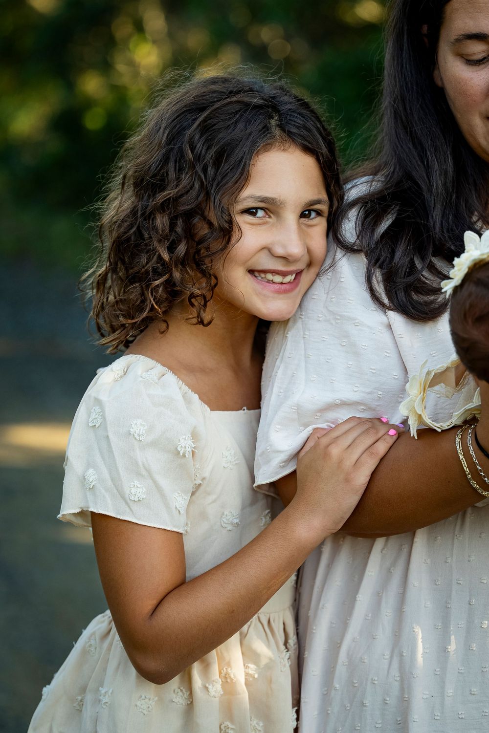 Happy young girl with dark curly hair in cream dress hugging adult outdoors.