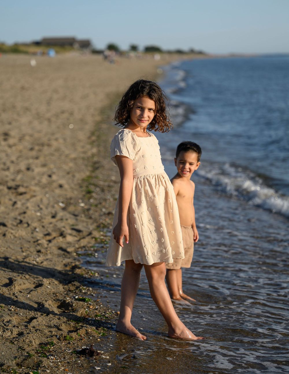 Two children standing barefoot at ocean shoreline on sunny beach day.