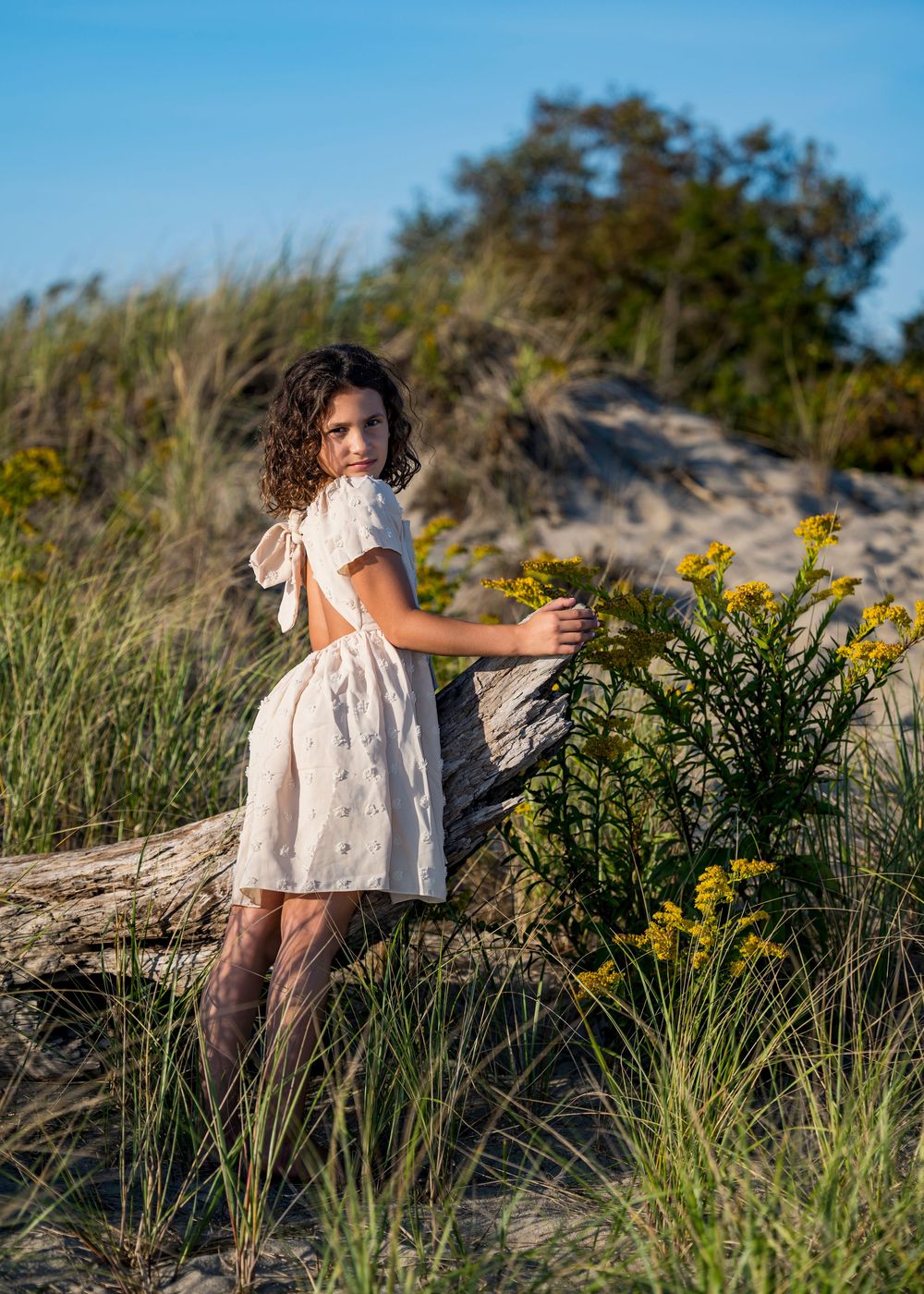 Young girl in cream dress standing by driftwood and wildflowers on beach.