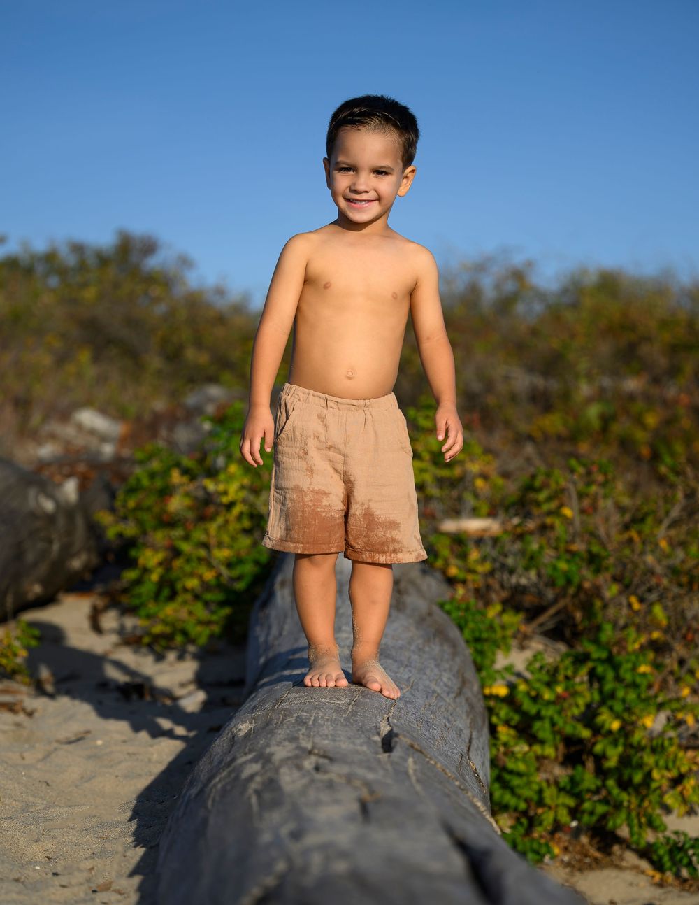 Young boy standing shirtless on driftwood log at sandy beach outdoors.