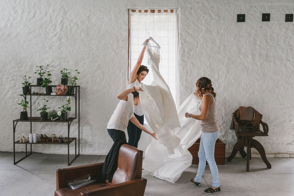 Bridesmaids unfold the bride's wedding dress by a window.