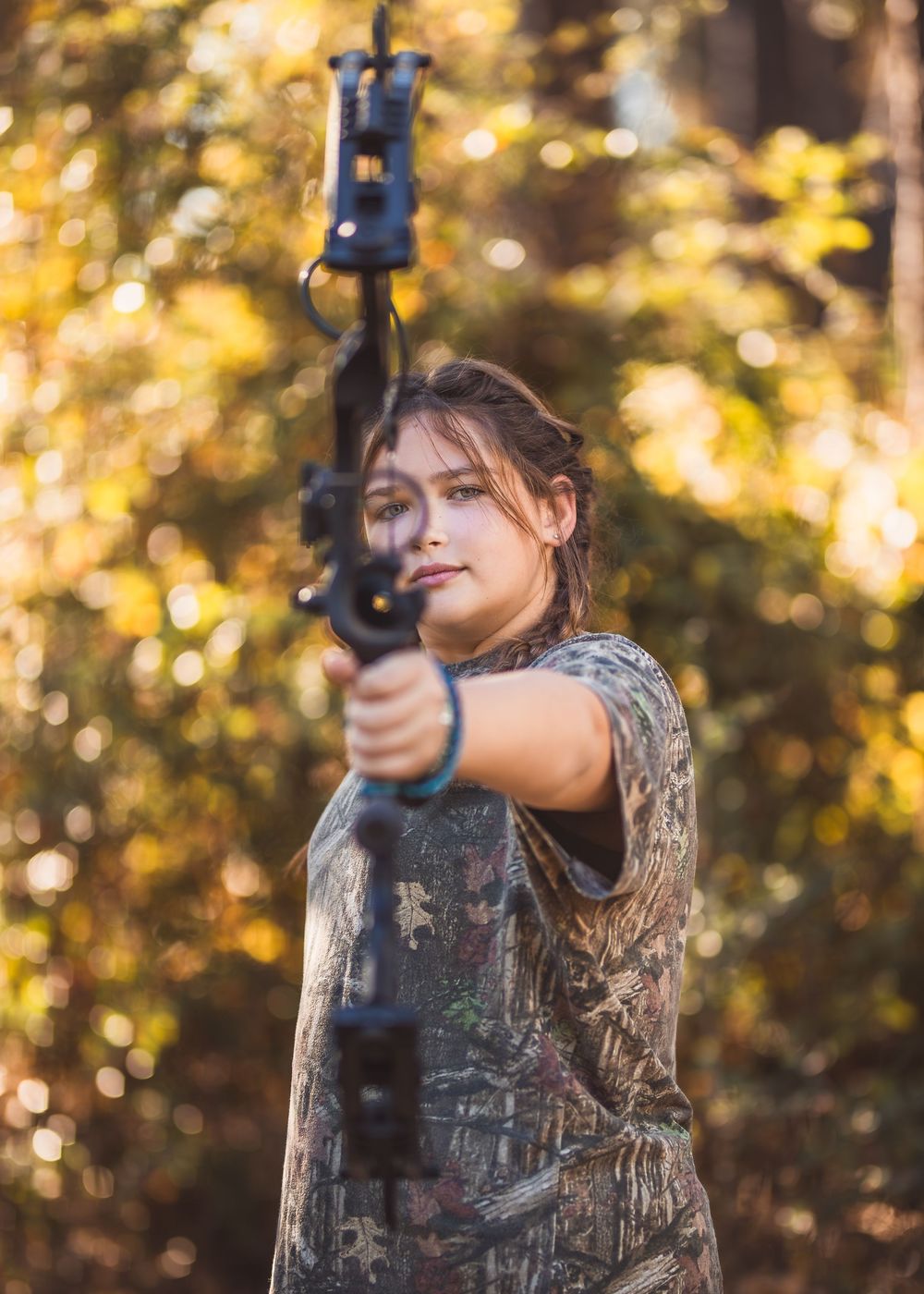 A girl in camouflage hunting gear aims a compound bow in an autumn forest setting with golden bokeh lighting for a portrait session.