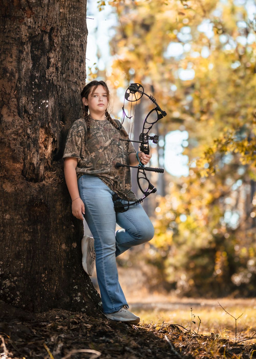Hunter in camouflage gear and blue jeans crouches near a tree trunk during autumn season with compound bow.