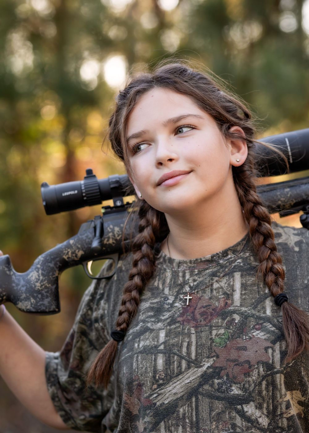 Young hunter in camouflage gear looking through rifle scope in outdoor woodland setting during golden hour.