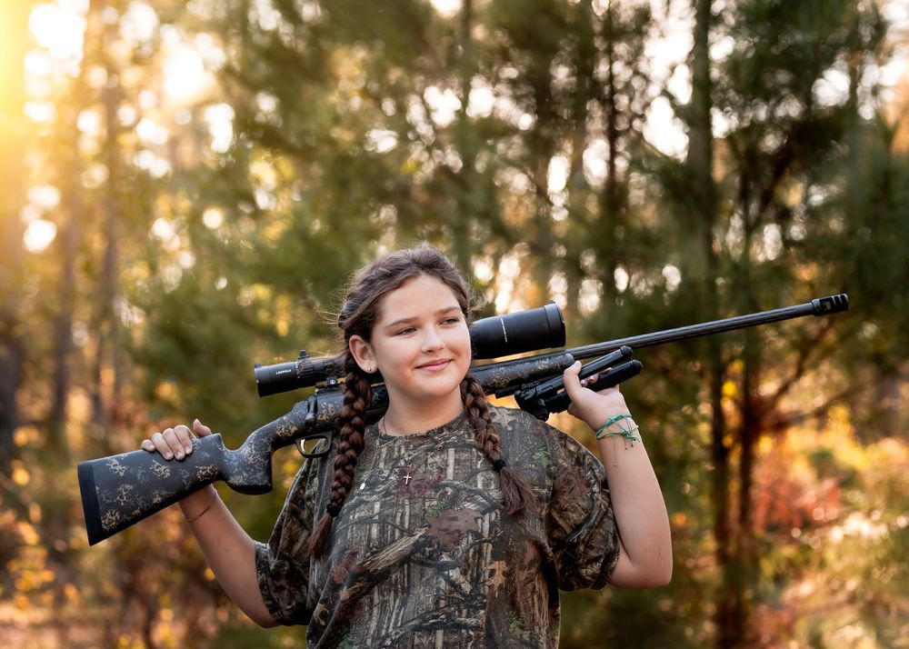 Young hunter in camouflage clothing holds a scoped rifle while standing in sunlit forest at golden hour posing for a photo session.