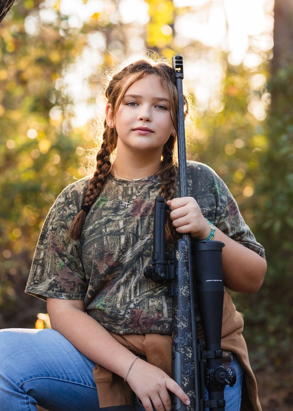 Young hunter in camouflage gear and jeans poses with a rifle in an autumn forest setting during golden hour.