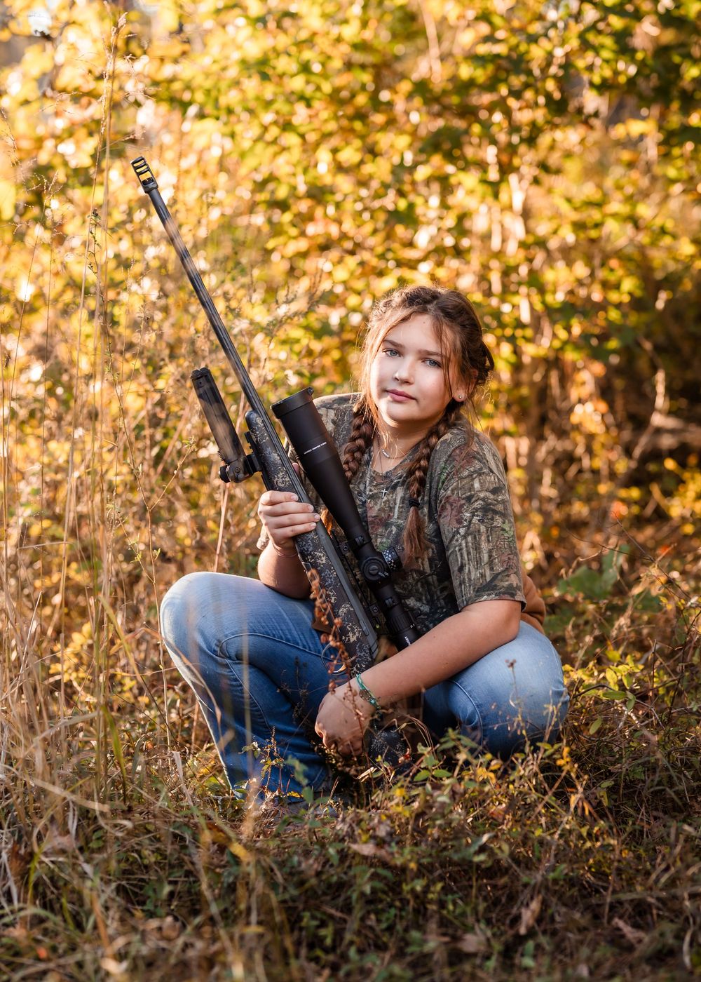 A girl in her camouflage hunting gear poses with a rifle in a golden autumn field during sunset.