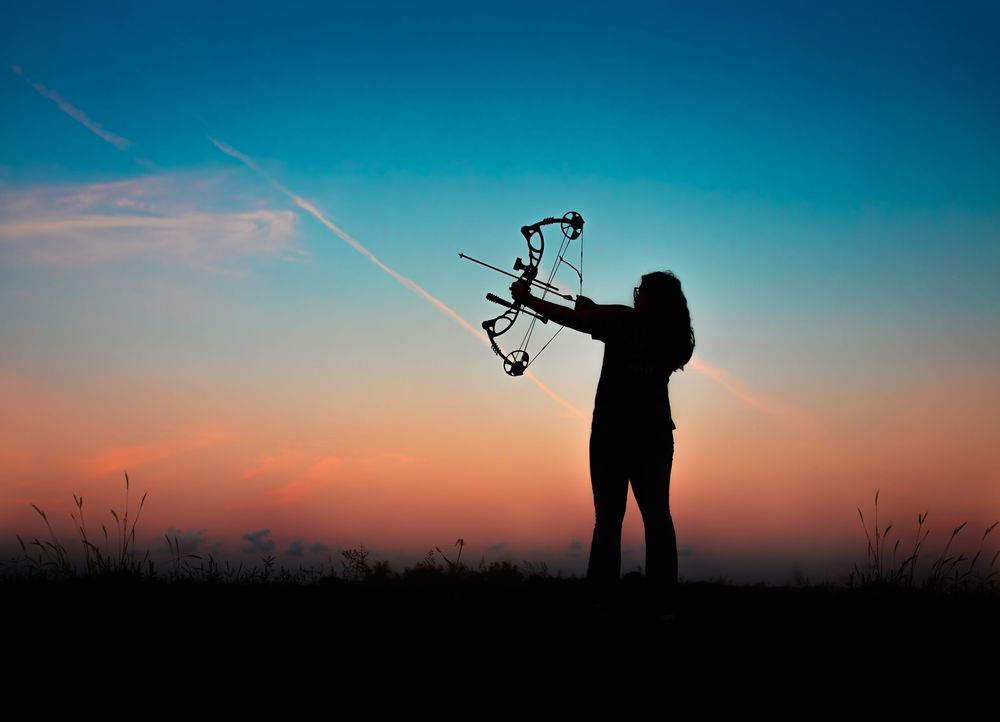 A silhouetted figure stands against a vibrant sunset sky while using a bow and arrow during a photoshoot.